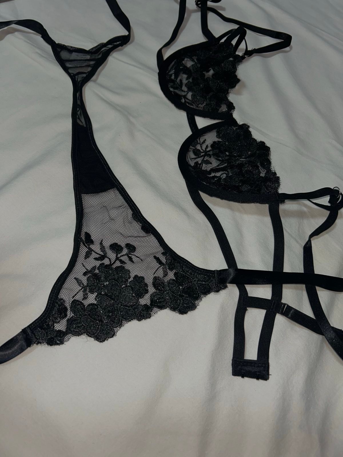 contact-img A sophisticated black lingerie set featuring sheer mesh panels, floral lace embroidery, and delicate strap details. The bra and matching bottom are displayed on a soft white fabric surface, highlighting the contrast between the dark lace and the light background while emphasizing the sensual and refined aesthetic of the piece.