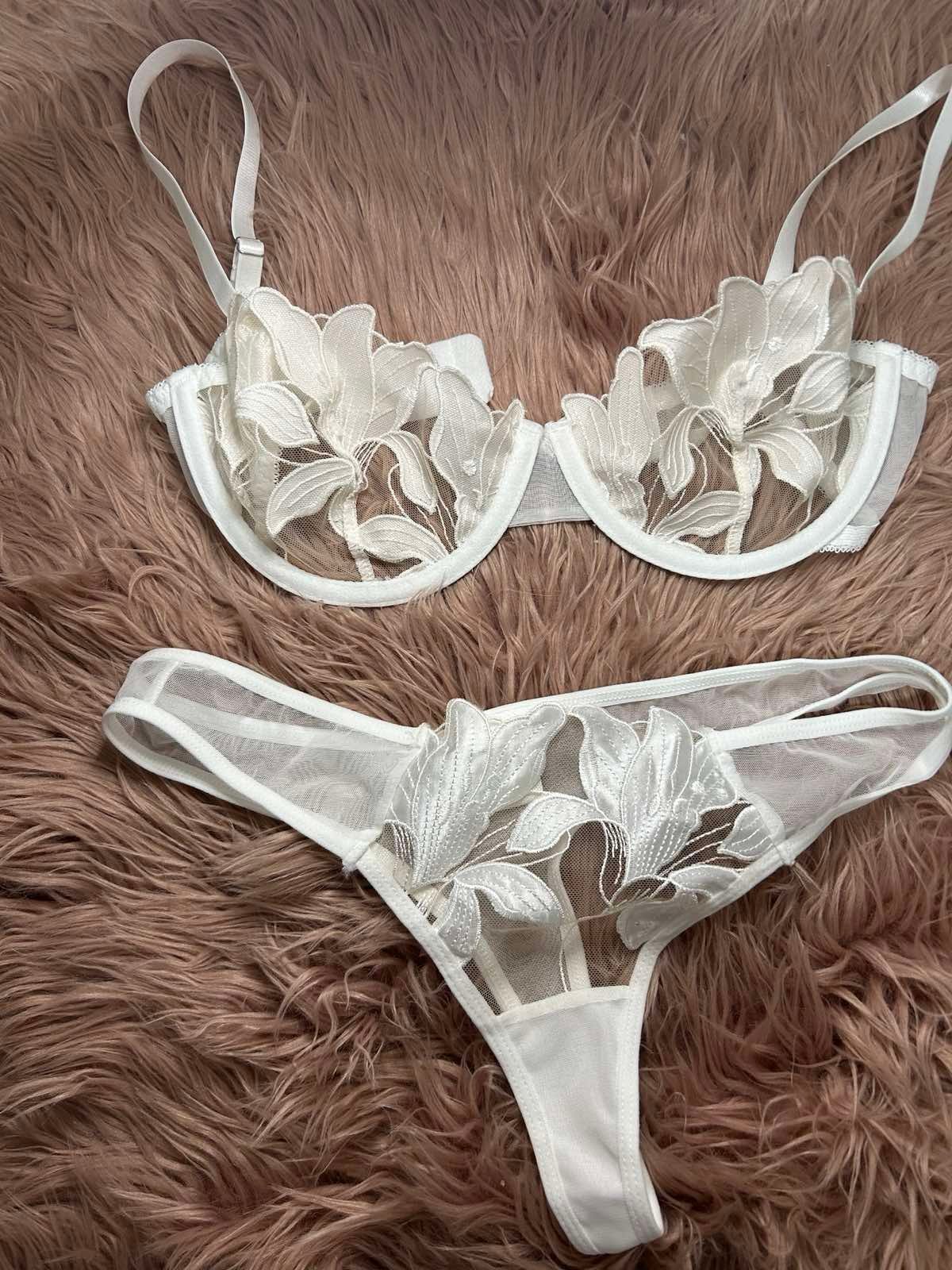 A delicate white lingerie set composed of a sheer underwire bra and matching thong, both decorated with large floral embroidery and soft mesh panels. The set is photographed flat on a blush faux-fur background, highlighting its feminine, refined, and romantic aesthetic.