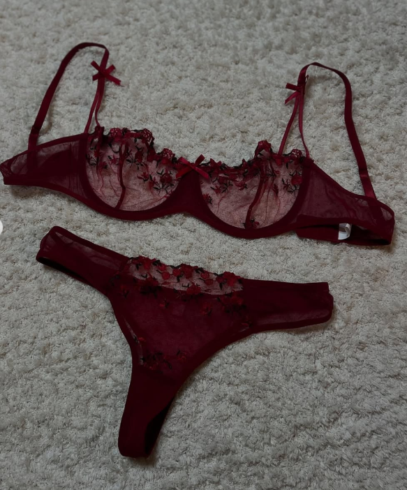 A romantic red lingerie set composed of a sheer underwire bra and matching thong, both decorated with delicate floral embroidery and fine mesh details. The set is displayed on a soft textured carpet, highlighting its feminine, sensual, and elegant aesthetic.